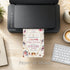 Printer on a desk with a printed card featuring floral designs and text, with 'Print at Home' text below.