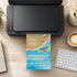 A black flatbed scanner sits on a wooden desk with a colorful beach-themed invitation card emerging from the bottom tray. The desk also includes a keyboard, a cup of latte, a small potted plant, and a notebook.