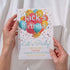 Hand holding a birthday party invitation with colorful balloons and text.