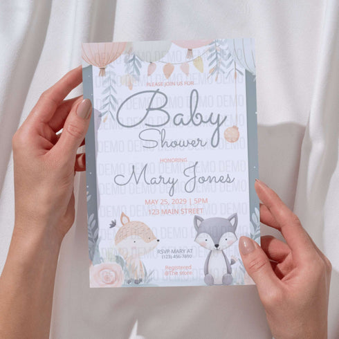 Baby shower invitation with fox and rabbit design held by hands against a neutral background