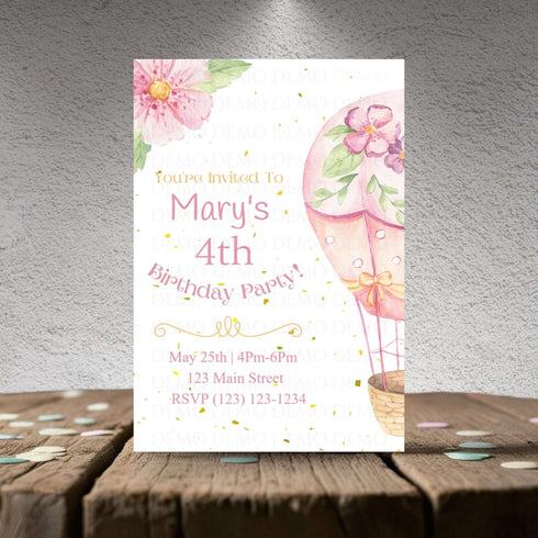 Birthday party invitation with pink hot air balloon and flowers on a wooden surface.