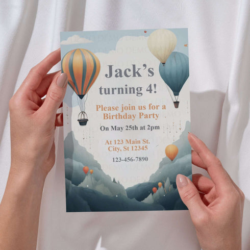 Birthday party invitation with hot air balloons held by hands against a white curtain background