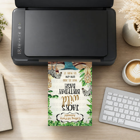 A colorful safari-themed birthday invitation card is shown printing from a black printer on a wooden desk, surrounded by a keyboard, coffee, and small plants.
