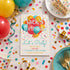 A birthday invitation card for 'Jack's Birthday!' is displayed on a decorated party table with balloons, confetti, plates, forks, napkins, and a slice of cake.