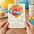 A person holds a birthday invitation card in front of a blurred party background. The card reads "Jack's Birthday!" with colorful balloon graphics, confetti, and event details.