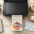A black printer sits on a wooden desk, printing a colorful birthday invitation card. The card features a playful balloon design in red, yellow, blue, and orange, with the text 'Jack's Birthday' and 'It's a Birthday!' in white.