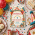 A circus-themed 7th birthday invitation card is displayed on a decorated birthday tablecloth surrounded by balloons, cupcakes, a slice of cake, a party hat, and confetti. The invitation features a vintage circus tent illustration with stars, balloons, a carousel horse, and readable event details for 'Jack is turning 7'.