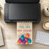 A colorful birthday invitation card with balloons is shown emerging from a black printer on a wooden desk, surrounded by a keyboard, coffee cup, notebook, and small plant.