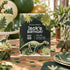 A dinosaur-themed birthday invitation card is displayed on a wooden table surrounded by matching party decorations, including plates, cups, balloons, and a decorated cake.