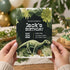 A person holds a dinosaur-themed birthday invitation card in front of a blurred festive background with balloons and plants. The card features a dark jungle design with a green dinosaur illustration and white event text reading 'Jack's Birthday' and 'May 22'.