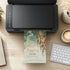 A black HP printer sits on a light wood desk, printing a decorative invitation card featuring a castle illustration and elegant script text. A keyboard, coffee cup, notebook, and small plant are visible around it in a styled workspace scene.