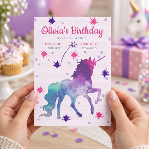A person holds a pastel unicorn birthday invitation card in front of a softly blurred party background with cupcakes, balloons, and a pink gift box. The card has a pink unicorn silhouette over a galaxy-style watercolor design in purple, blue, and teal, with the text 'Olivia's Birthday' and party details.