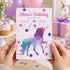 A person holds a pastel unicorn birthday invitation card in front of a softly blurred party background with cupcakes, balloons, and a pink gift box. The card has a pink unicorn silhouette over a galaxy-style watercolor design in purple, blue, and teal, with the text 'Olivia's Birthday' and party details.
