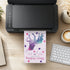 A black printer sits on a wooden desk with a colorful birthday invitation card emerging from the front. The card features a purple and blue watercolor silhouette of a person with stars and the text 'Olivia's Birthday'.