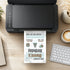 A black desktop printer on a light wood desk, printing a personalized birthday invitation card. The card features a white background with blue wavy lines, black text, and small illustrations of a pair of boots, a patterned top hat, and a silver teapot.