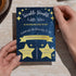 A person holds a navy blue and gold 'Twinkle, Twinkle Little Star' baby shower invitation card with hands against a light stone countertop background.
