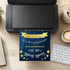 A custom printed navy blue and gold wedding invitation card is shown emerging from a black printer on a wooden desk, surrounded by a keyboard, coffee cup, and other office props.