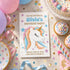 A unicorn birthday party invitation card is displayed in a styled flat-lay scene with balloons, cupcakes, confetti, ribbons, and a party hat.