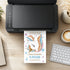 A colorful unicorn birthday party invitation card is shown emerging from a black printer on a wooden desk, surrounded by a keyboard, coffee cup, and other desk accessories.