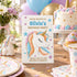A unicorn-themed birthday invitation card displayed on a decorated party table with cupcakes, plates, balloons, and a small unicorn figurine.