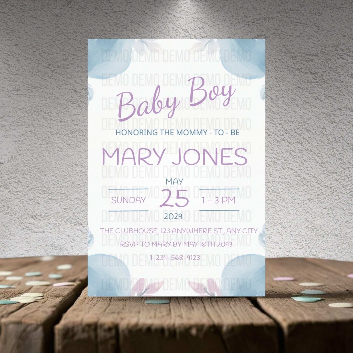 Baby shower invitation card for a baby boy on a wooden surface with a textured gray wall background.