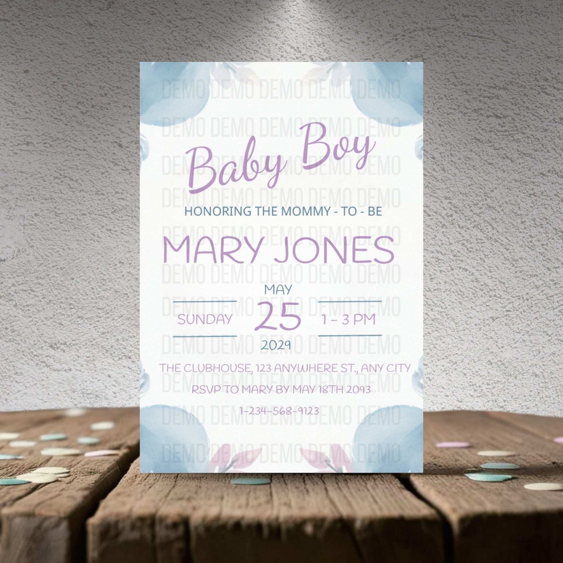 Baby shower invitation card for a baby boy on a wooden surface with a textured gray wall background.