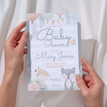 Baby shower invitation with fox and rabbit design held by hands against a neutral background