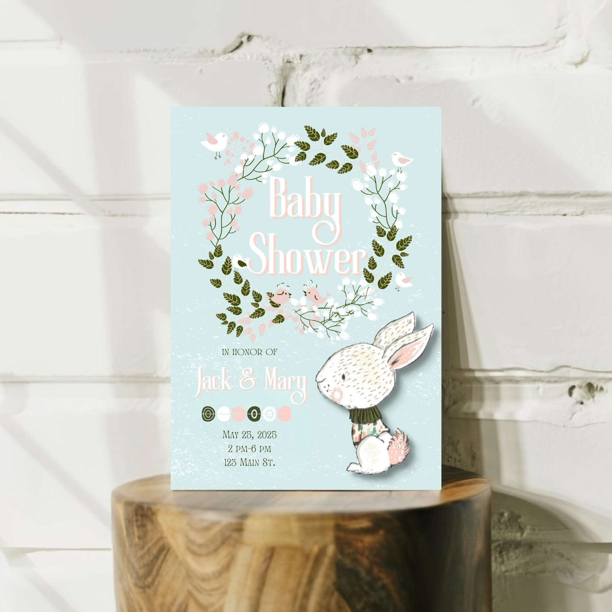 A personalized floral wreath baby shower invitation with a cartoon image of a bunny on it, set against a wooden background.
