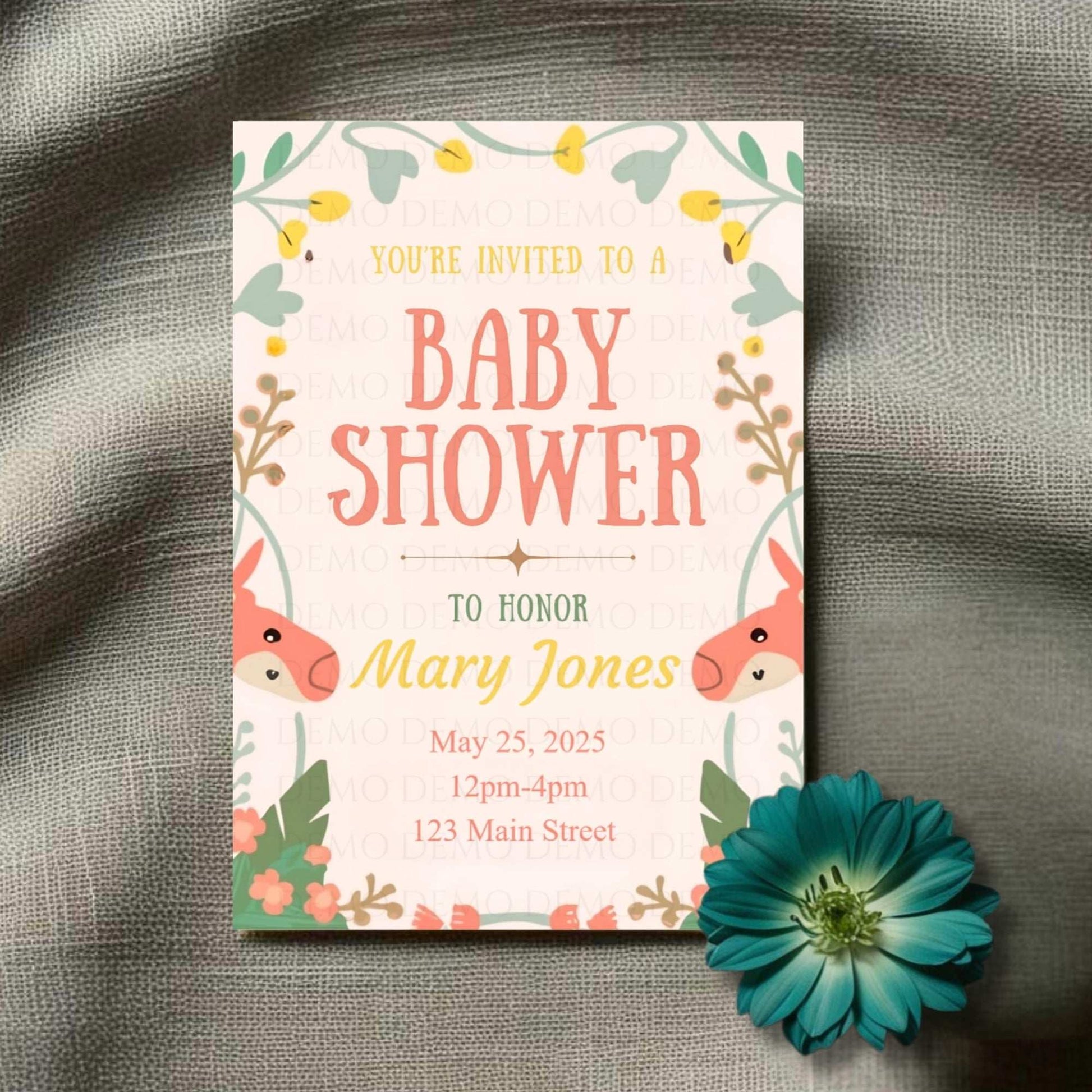 A baby shower invitation card with a floral border and cartoon animals on it, laid on a textured fabric background with a blue flower beside it.