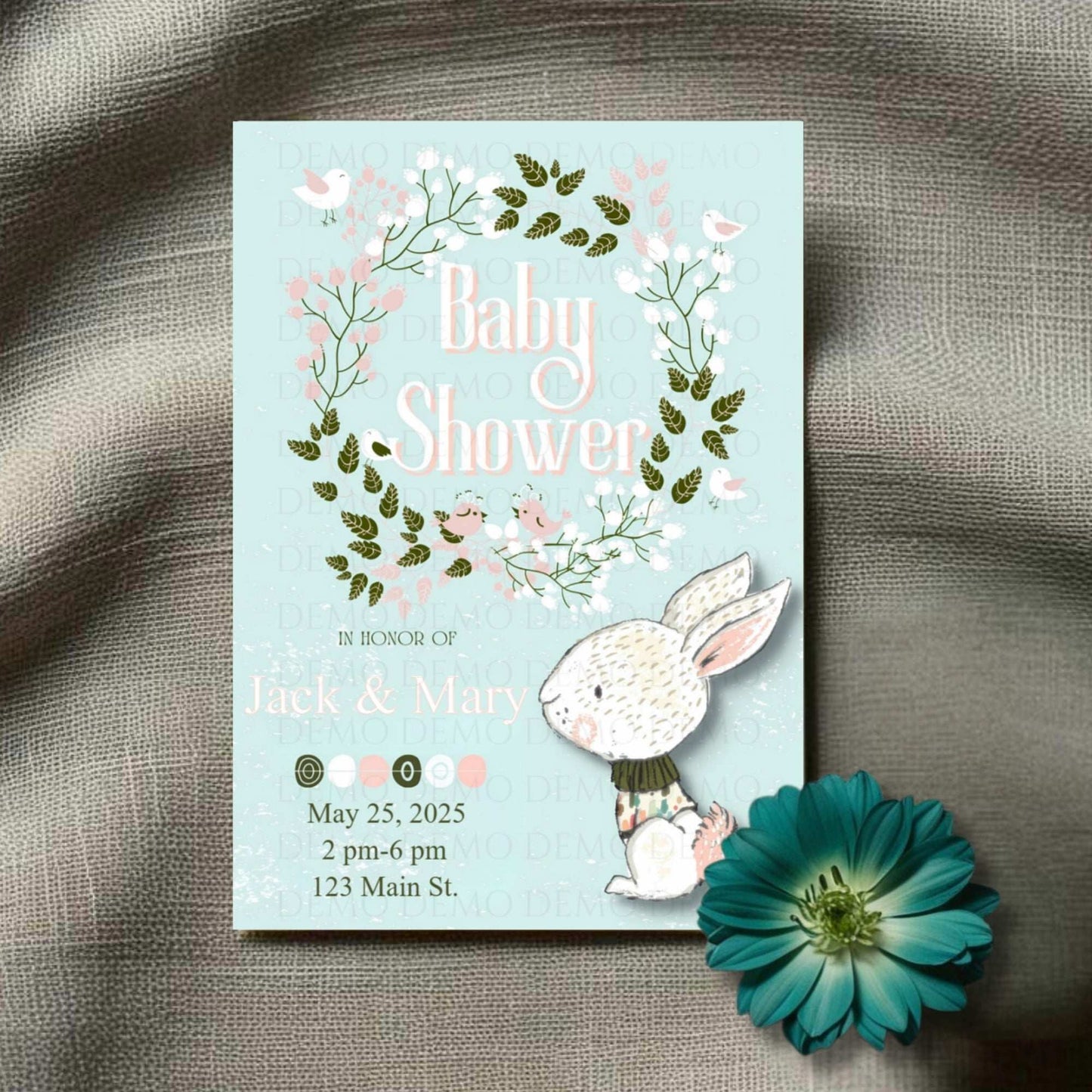 A personalized floral wreath baby shower invitation with an illustration of a bunny and flowers on a card, placed on a fabric surface with a flower decoration.