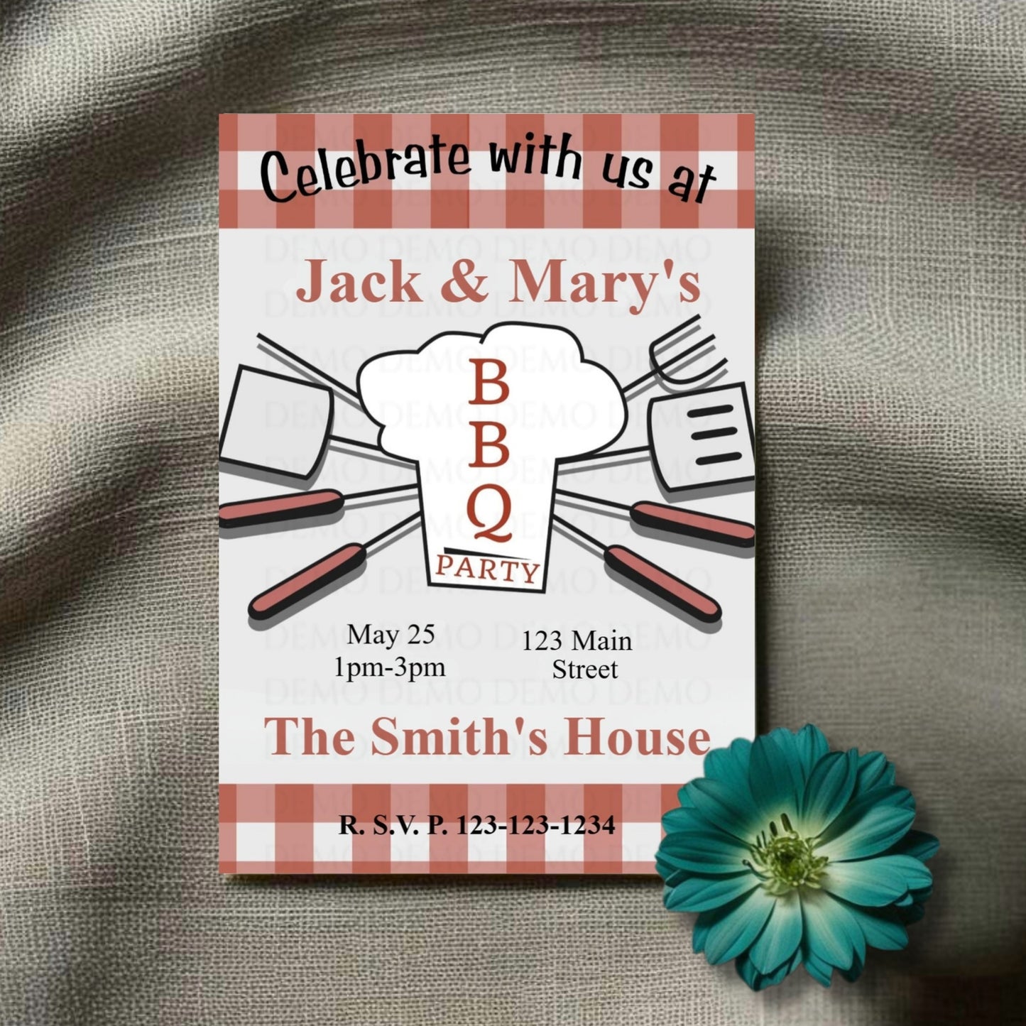 A personalized housewarming invitation with a plaid pattern and BBQ theme laid out on a fabric surface with a decorative flower beside it.