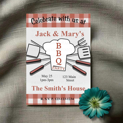 A personalized housewarming invitation with a plaid pattern and BBQ theme laid out on a fabric surface with a decorative flower beside it.