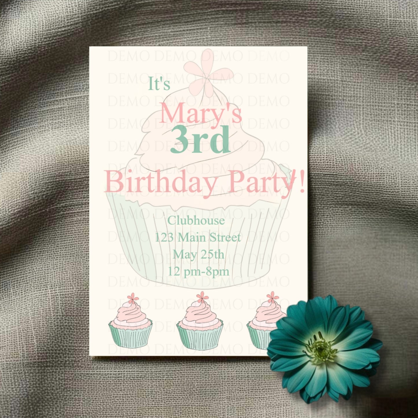 A personalized green-pink cupcake invitation card for a 3rd birthday party laid on a grey surface with a blue flower decoration.