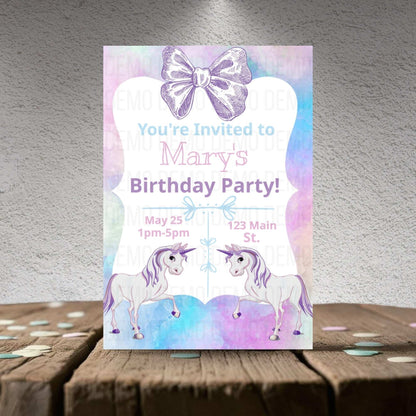 Birthday party invitation with unicorns and a bow on a wooden surface.