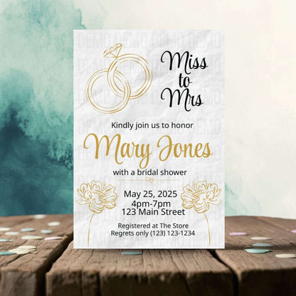 Bridal shower invitation with gold rings and floral design on a wooden surface with a watercolor background.
