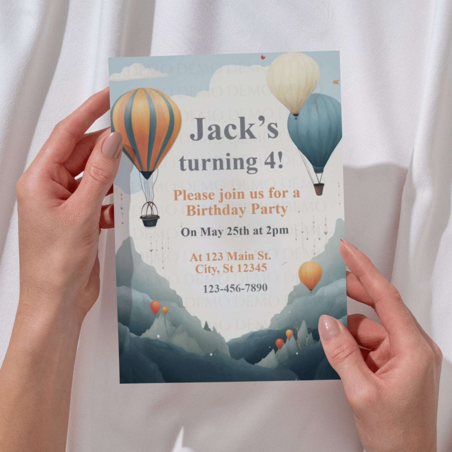 Birthday party invitation with hot air balloons held by hands against a white curtain background