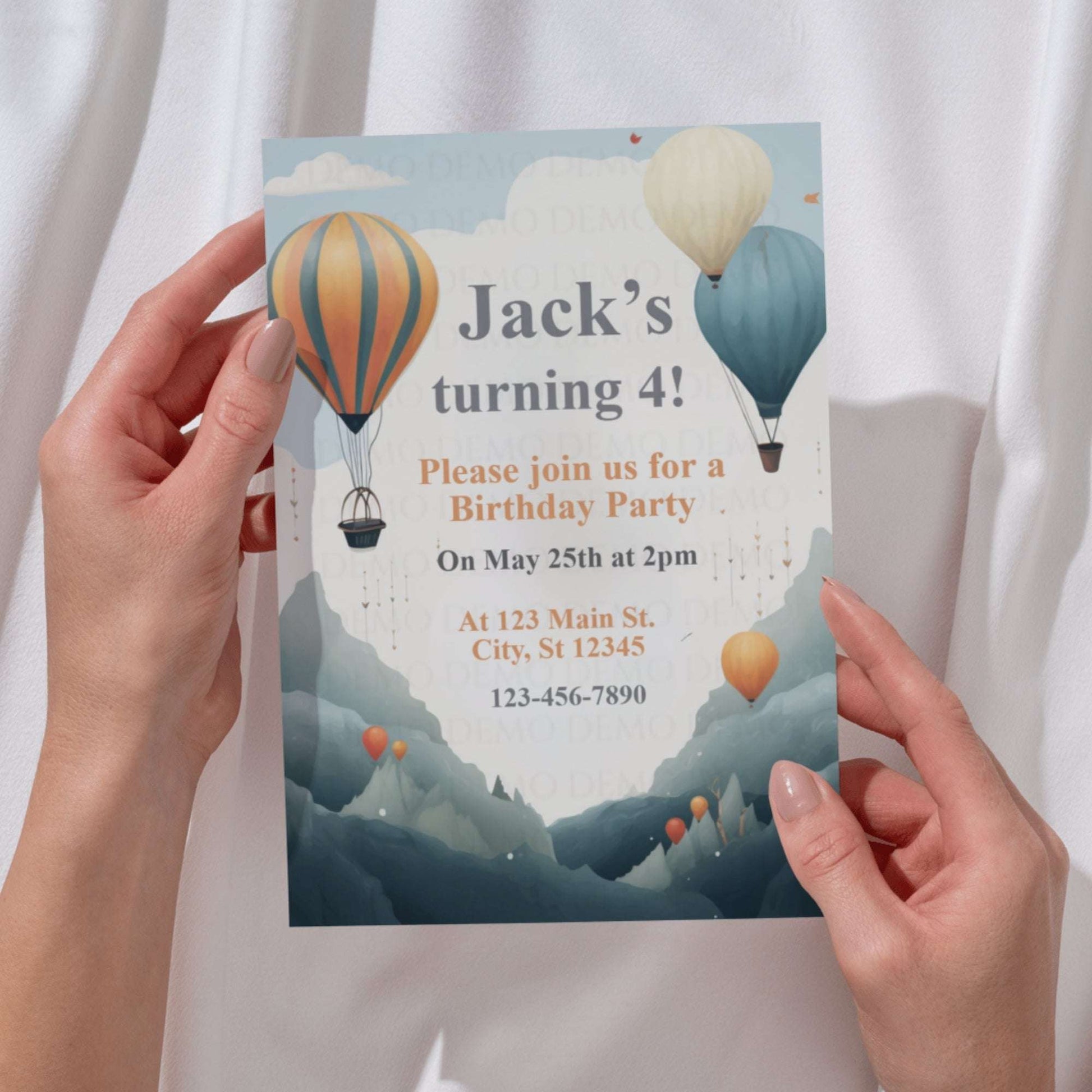 Birthday party invitation with hot air balloons held by hands against a white curtain background